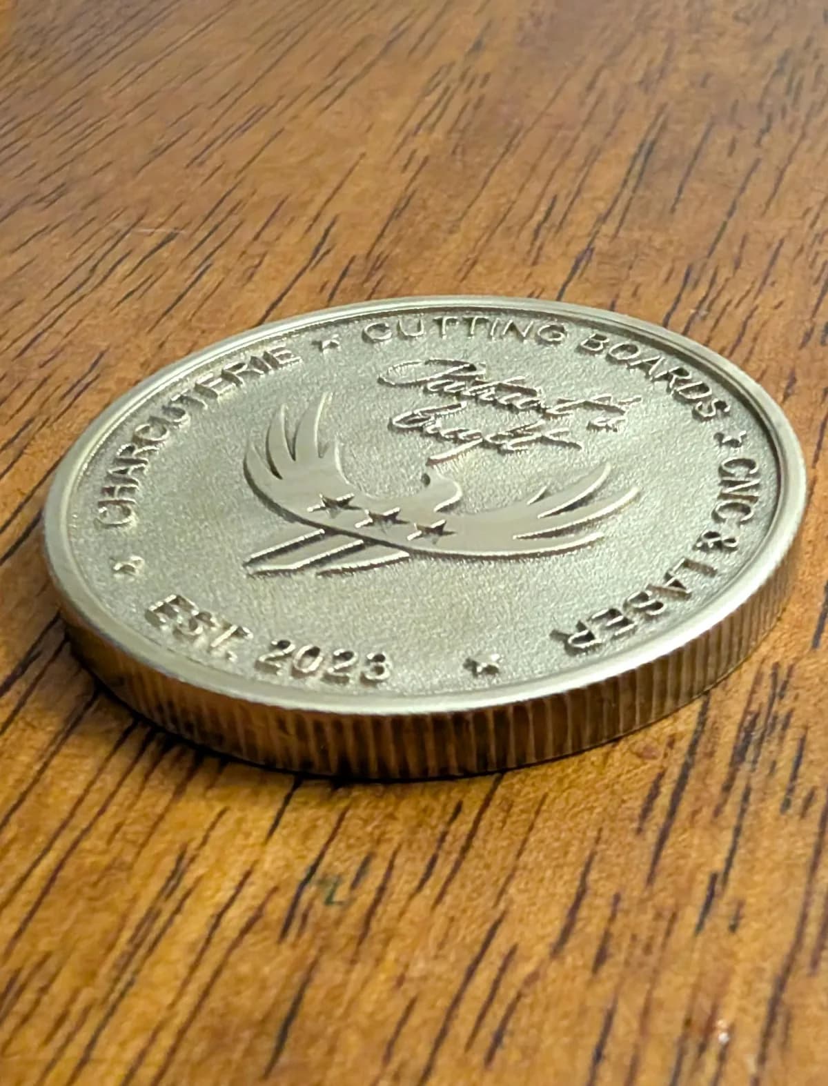 Brass round medallion laser engraved with Patriot's Craft logo and circle text surrounding logo near the edge of medallion.